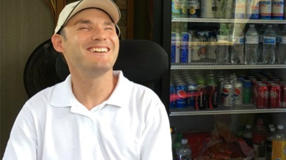 Smiling young man in white shirt and hat sitting in front of beverage refrigerator