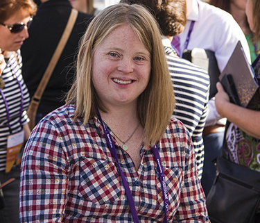 Woman with blonde hair and a plaid shirt smiling at camera during The Arc National Convention, people mingling in background