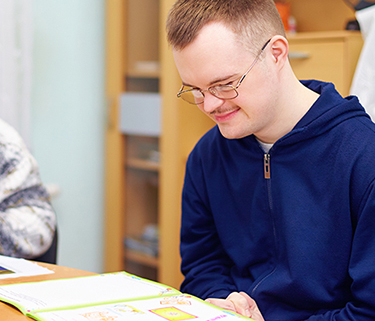 A young man wearing glasses smiles while sitting at a desk and reading a booklet
