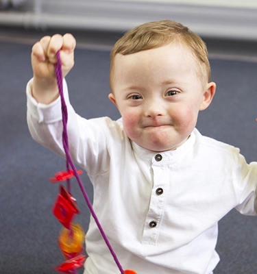Toddler with Down syndrome sits on a floor and proudly holds up his toy necklace