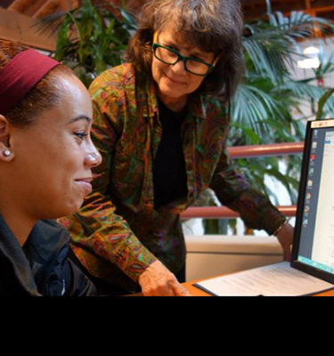 A woman smiles as she looks at a computer, while another woman stands next to her smiling