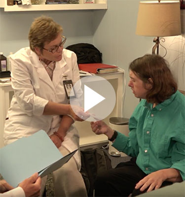 Two women, a doctor and a patient, sit in an appointment. The doctor is passing a paper to the patient.